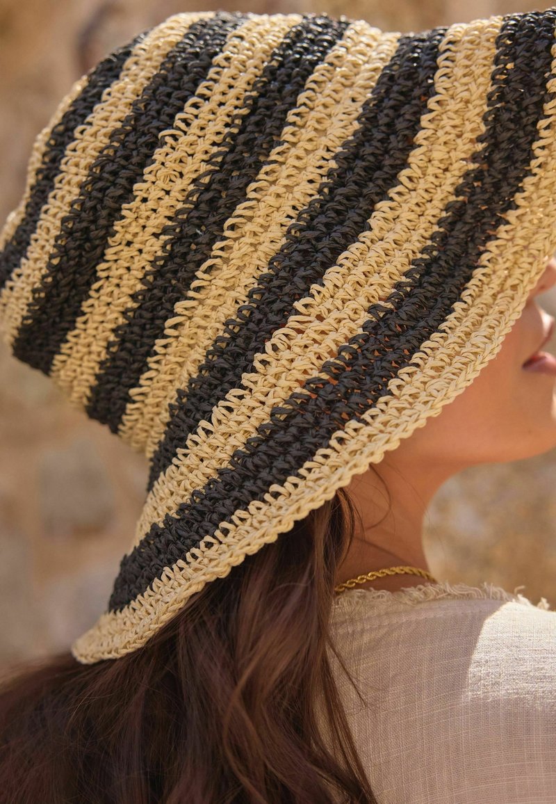 Femme portant un chapeau crocheté beige et noir à larges rayures, avec de longs cheveux bruns et un collier en or visible à la nuque.