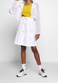 Vita lätta shorts med sidofickor, kombinerat med en gul figurnära topp och en genomskinlig vit kort jacka. Svart och vita sneakers kompletterar outfiten.