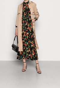 Žena v béžovém trenčkotě přes černé květované midi šaty, držící černou kabelku a v černých lodičkách se zapínáním kolem kotníku.