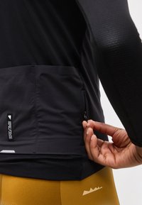 Χέρι που ρυθμίζει μικρό φερμουάρ στο κάτω μέρος της πίσω πλευράς μαύρου αθλητικού μπουφάν που φοριέται πάνω από μουσταρδί κίτρινες βερμούδες.