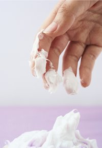 Biała kremowa masa rozprowadzona na opuszkach palców, gładka konsystencja, lekko błyszczące wykończenie, na jasnym tle, z delikatnym fioletowym podkładem.