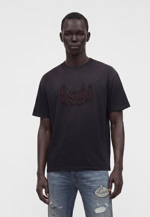 Man draagt een zwart T-shirt met rode geborduurde tekst en gescheurde blauwe spijkerbroek met merkpatch, staand tegen een effen lichte achtergrond.