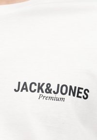 Άσπρο ύφασμα με μαύρο κείμενο «JACK & JONES Premium» τυπωμένο στην περιοχή του στήθους ενός πουκάμισου.