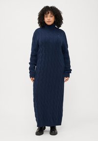 Frau mit lockigem Haar, die ein langes, marineblaues, gestricktes Kleid mit Rollkragen und Rippbündchen trägt, steht vor einem weißen Hintergrund.