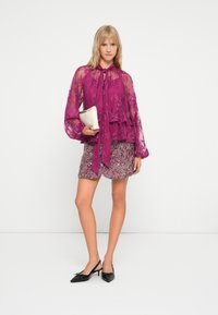 Bluză fuchsia transparentă cu guler legat, design cu volane și mâneci lungi, asociată cu o fustă scurtă texturată și cu model și pantofi cu toc negri.