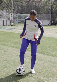 Atuendo atlético blanco y morado, con un logo del Olympique Lyonnais, combinado con zapatillas blancas y un balón de fútbol en blanco y negro sobre hierba.
