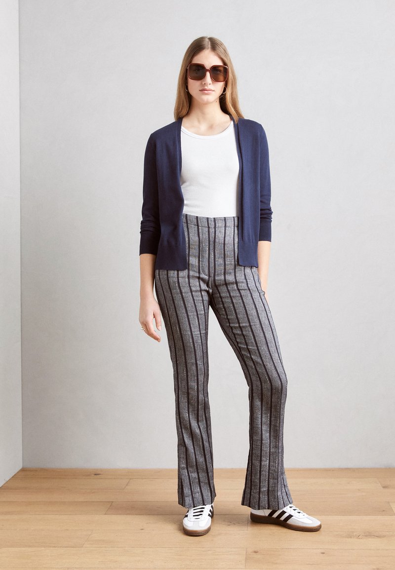 Cardigan navy, top blanc, pantalon évasé rayé gris, baskets blanches avec accents noirs, grandes lunettes de soleil brunes. Tissu lisse, design ajusté.