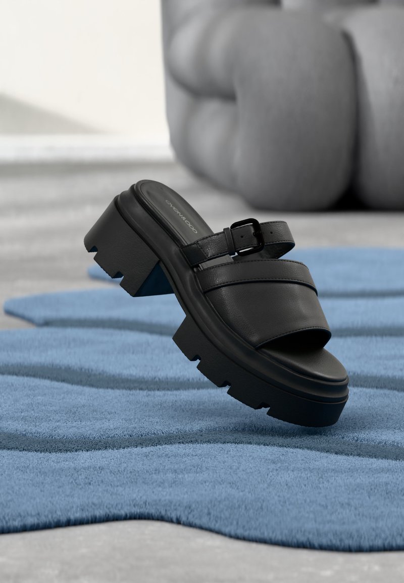 Sandale noire à talon carré avec bride à boucle flottant au-dessus d'un tapis bleu texturé, avec des jambes floues pliées en arrière-plan.