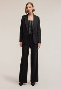 Zwart getailleerd colbert met geribde revers, gecombineerd met een strakke zwarte corset en hoog getailleerde zwarte broek. Overal een gladde textuur.