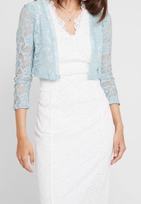 Lichtblauw kanten bolero met driekwart mouwen over een witte kanten jurk met een golvende halslijn en een aansluitende snit.