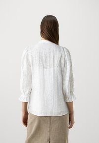 Vit långärmad blus i texturerad tyg, rund krage och sammansatta puffärmar. Subtilt lövmönster framhäver designen.