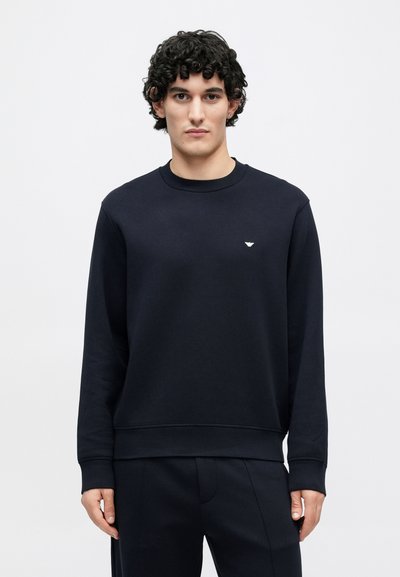 Námornícka modrá mikina z mäkkého materiálu, s okrúhlym výstrihom, dlhými rukávmi a malým logom na hrudi. Voľnejší strih s rebrovanými manžetami.