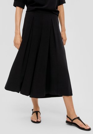 Frau trägt einen schwarzen plissierten Midirock und schwarze Zehensandalen, steht vor einem schlichten Hintergrund.