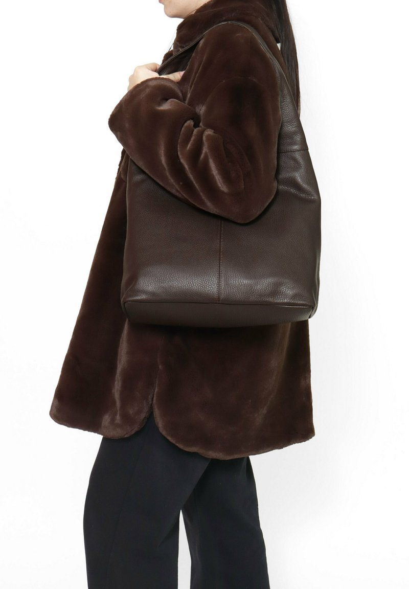 Borsa tote in pelle marrone con finitura testurizzata, caratterizzata da un ampio corpo e da una robusta tracolla, portata insieme a un cappotto marrone morbido.