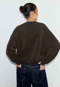 Rjava prevelika jopa z spuščenimi rameni in rebrastim izrezom, združena z modrimi denim jeans hlačami z vidnimi šivi in žepi.