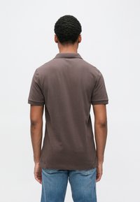 Homem com cabelo curto e encaracolado, vestindo uma camisa polo castanha e jeans azuis, em pé de frente para um fundo branco simples.