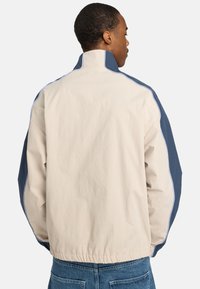 Lightweight jacket featuring a cream body with navy blue sleeves, elastic cuffs, and subtle blue piping along the seams.