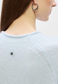 Světle modrý pletený svetr s kulatým výstřihem, který má na zadní straně malý černý dekorativní knoflík a texturovaný vzor stehu.