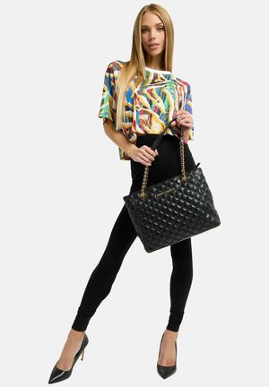 Schwarze gesteppte Tote-Bag mit goldenen Kettenriemen, die in der Hand eines Models gehalten wird, kombiniert mit einem bunten, gemusterten Kurzarmoberteil und schwarzen Leggings.