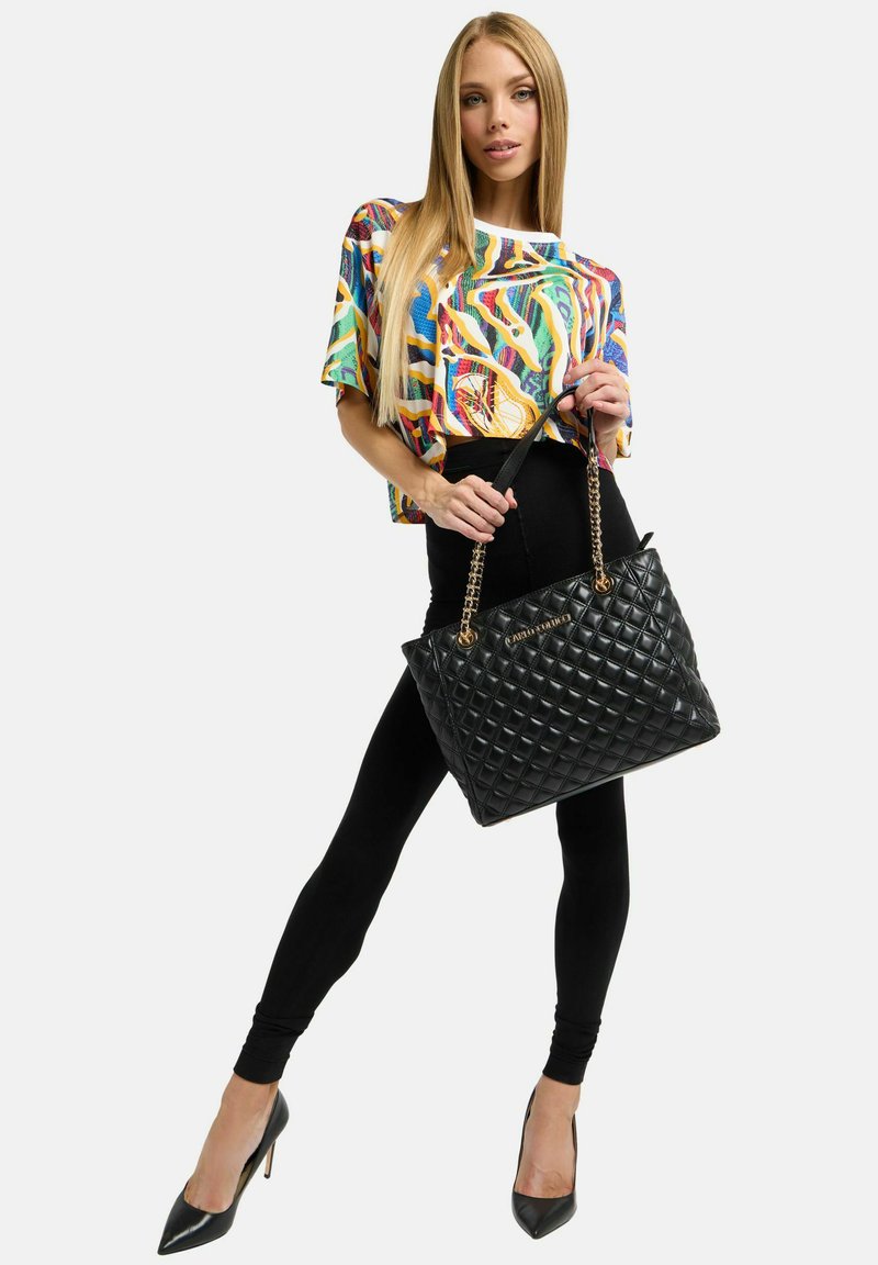 Schwarze gesteppte Tote-Bag mit goldenen Kettenriemen, die in der Hand eines Models gehalten wird, kombiniert mit einem bunten, gemusterten Kurzarmoberteil und schwarzen Leggings.