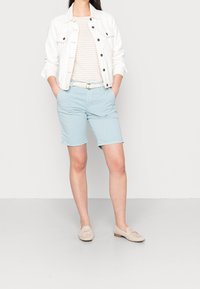 Biała kurtka dżinsowa narzucona na beżowo-białą pasiastą koszulę, zestawiona z jasnoniebieskimi szortami i beżowymi balerinami. Casualowy strój w neutralnych tonach.