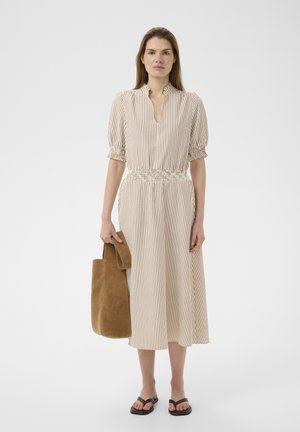 Kobieta w midi sukience z pionowymi beżowo-białymi paskami, trzymająca brązową zamszową torbę na ramię, w czarnych sandałach, stojąca na jednolitym tle.