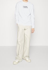 Person trägt einen weißen Sweatshirt mit dem Aufdruck "KARL LAGERFELD", beigen weit geschnittenen Hosen und weißen Plateausneakern, stehend vor einem schlichten Hintergrund.