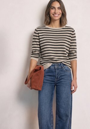 Femme souriante portant un pull rayé beige et noir, un jean bleu, tenant un sac à main couleur rouille dans sa main droite sur un fond uni.