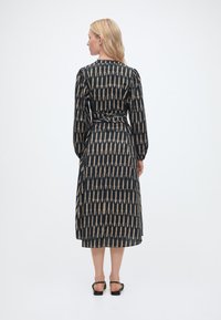 Γυναίκα με ξανθά μαλλιά στέκεται προς τα πίσω φορώντας μαύρο midi φόρεμα με μπεζ γεωμετρικό σχέδιο και μαύρα σανδάλια με ανοιχτή φτέρνα.