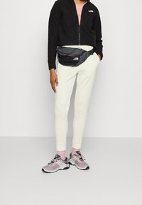 Giacca nera corta con zip, maglietta rosa chiaro, pantaloni tuta crema, marsupio nero e scarpe sportive grigie-rosa con dettagli neri.