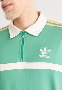 Grön zippar polo t-shirt med vit krage, med vit horisontell rand och broderad Adidas-logotyp; har gula accentrån på ärmarna.