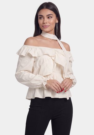 Frau mit langen dunklen Haaren, die eine cremefarbene Carmenshirt mit Rüschen und schwarze Hose trägt, Hände vor dem Körper verschränkt.