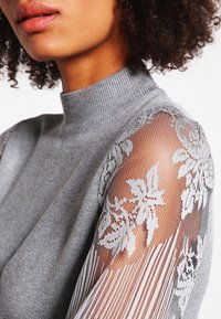 Grauer Pullover mit hohem Kragen und körpernahem Schnitt; durchsichtige Spitzenärmel mit floralen Mustern und geripptem Textur an den Schultern.