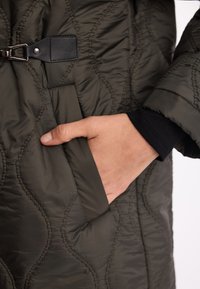 ciemnozielona pikowana kurtka o teksturowanej powierzchni, z boczną kieszenią oraz czarnym skórzanym akcentem z srebrną klamrą.