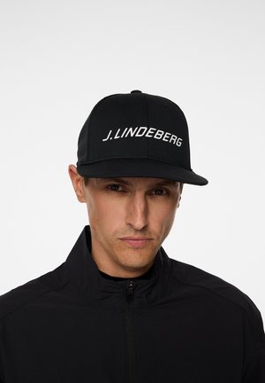 Czarna czapka z płaskim daszkiem i białym napisem "J. Lindeberg" z przodu. Tkanina jest teksturowana i strukturalna, co dobrze współgra z czarną kurtką poniżej.