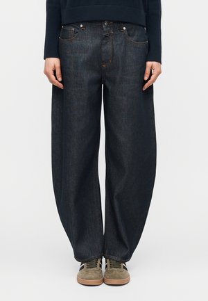 Άτομο που φοράει άνετο σκούρο τζιν παντελόνι, σκουρόχρωμο μακρυμάνικο μπλουζάκι και μπεζ αθλητικά παπούτσια με καφέ σόλες, στέκεται μπροστά σε απλό φόντο.