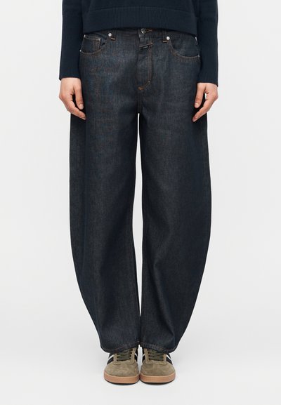 Person trägt locker sitzende dunkle Jeans, ein dunkles langärmeliges Oberteil und beige Sneaker mit braunen Sohlen, steht vor einem schlichten Hintergrund.