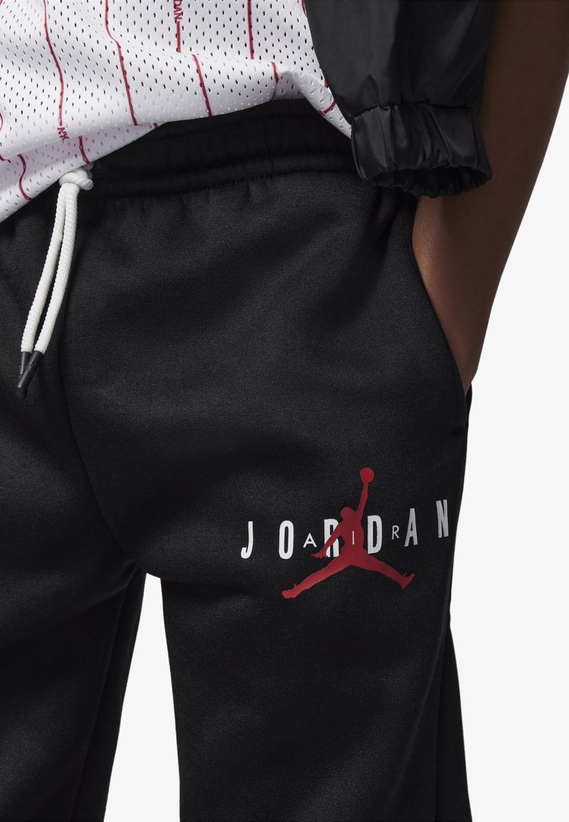 Joggers de sport noirs en tissu lisse avec un cordon blanc. Dotés d'un logo "JORDAN" rouge et d'une illustration en rouge vif.