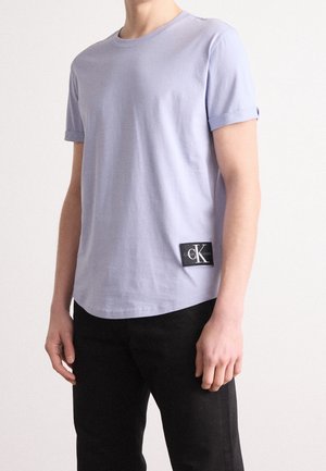 Lila Baumwoll-T-Shirt mit kurzen Ärmeln, Rundhalsausschnitt und einem schwarzen Logo-Patch auf der unteren linken Seite. Kombiniert mit schwarzen Hosen.