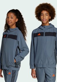 Veste de sport grise avec capuche, dotée de bandes noires et du logo orange Adidas. Le tissu semble lisse avec des poignets et une taille élastiques.