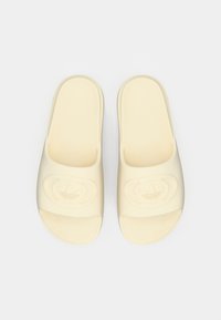 Jasne klapki z gumy, z wytłoczonym logo na pasku i otwartym przodem dla łatwego zakładania.