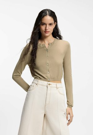 Kobieta z długimi ciemnymi włosami, ubrana w beżowy zapinany na guziki kardigan i kremowe szerokie spodnie z wysokim stanem na jednolitym białym tle.