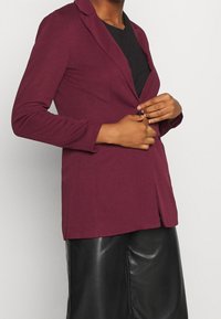 Person som knäpper en vinröd kavaj över en svart skjorta, matchad med svarta läderbyxor, mot en ljus enfärgad bakgrund.