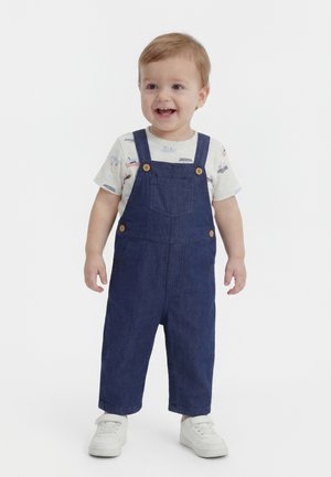 Smårolling med lysebrunt hår som har på seg en hvit skjorte med båttrykk, mørkeblå hengebukser og hvite joggesko, smilende og stående.