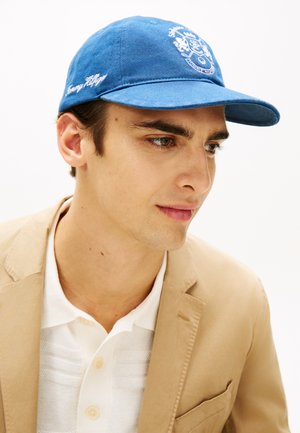 Ung mand iført en blå Tommy Hilfiger-kasket, beige blazer og hvid skjorte med knapper, kigger let mod højre mod en hvid baggrund.