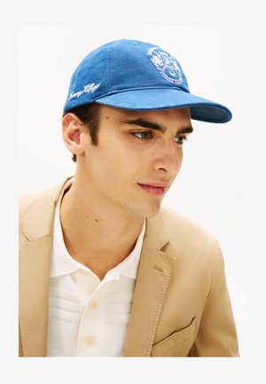 Ung mann som bærer en blå Tommy Hilfiger-caps, beige blazer og hvit skjorte med knapper, som ser lett mot høyre mot en hvit bakgrunn.