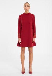 Rotes Kleid mit hohem Kragen und langen Ärmeln, verziert mit Spitzenelementen an den Bündchen. Ausgestellte Silhouette, aus glattem Stoff gefertigt.
