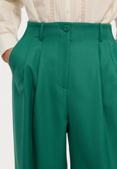 Groene op maat gemaakte broeken met een visgraatstructuur, voorzien van voorplooien, zijzakken en een enkele knoopsluiting in de taille.