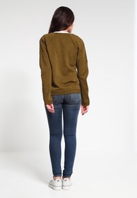 Sudadera marrón texturizada con mangas largas, cuello redondo y un escote blanco en contraste. Llevada con jeans ajustados de color azul claro y zapatillas blancas.