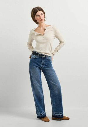 Jeune femme portant un t-shirt henley beige à manches longues, jean bleu taille haute à jambes larges, ceinture noire et mocassins marron, debout les mains sur les hanches.